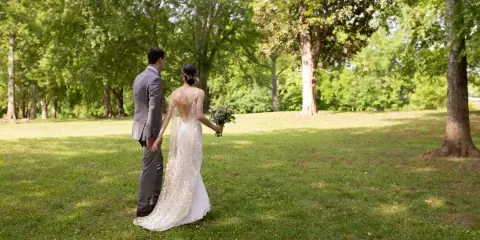 A bride and groom standing in a grassy field with trees in the background, holding hands and a bouquet.