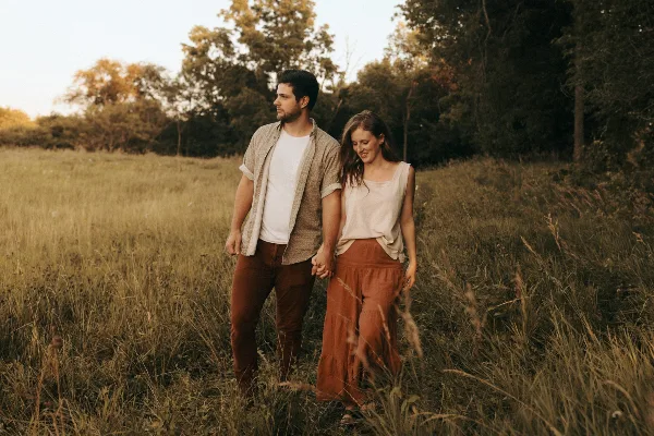 A couple holding hands and walking through a grassy field at sunset.
