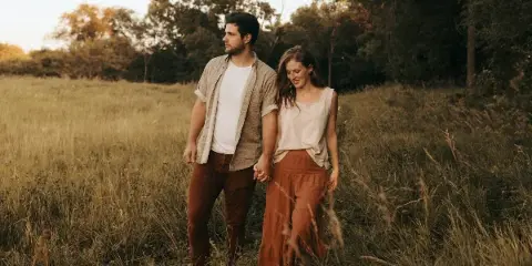 A couple holding hands and walking through a grassy field at sunset.