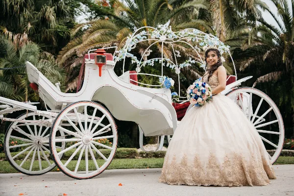 A bride in a white wedding dress holding a bouquet poses next to a white carriage decorated with flowers.