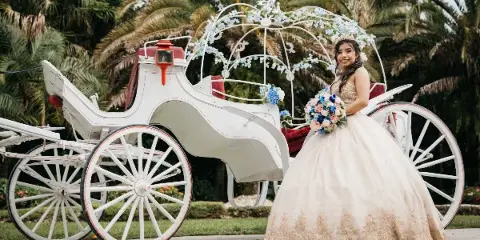 A bride in a white wedding dress holding a bouquet poses next to a white carriage decorated with flowers.