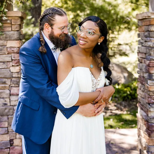 A groom in a blue suit and a bride in a white dress embracing outdoors near a stone archway.