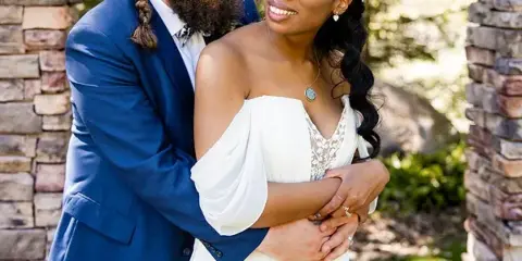 A groom in a blue suit and a bride in a white dress embracing outdoors near a stone archway.