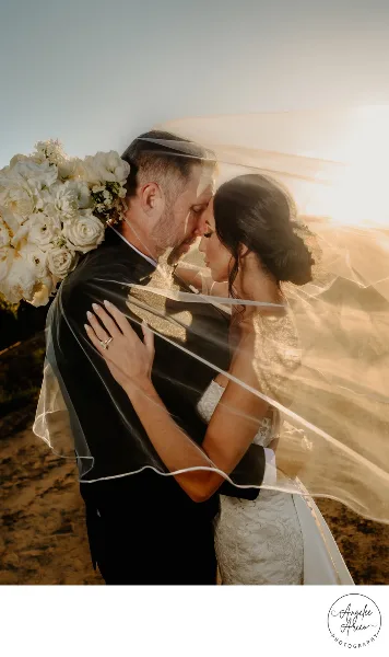 A bride and groom embracing on a beach at sunset, with the bride holding a large white bouquet and wearing a veil.