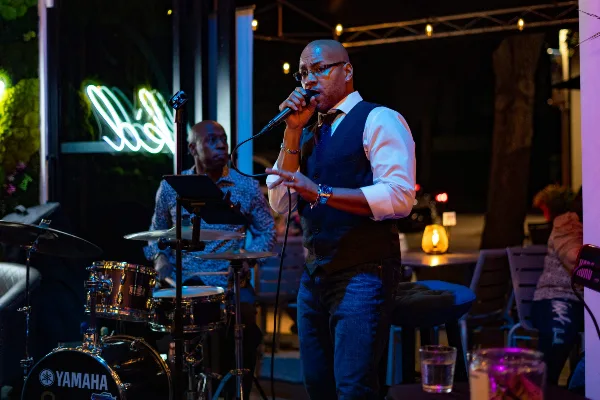 A man in a vest and tie singing into a microphone on stage with a drummer in the background.