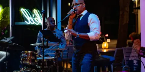 A man in a vest and tie singing into a microphone on stage with a drummer in the background.