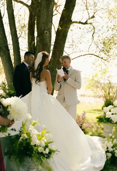 A bride and groom standing at an outdoor altar with an officiant during a wedding ceremony, surrounded by white flowers and trees.