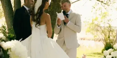 A bride and groom standing at an outdoor altar with an officiant during a wedding ceremony, surrounded by white flowers and trees.