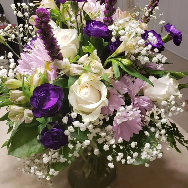 A floral arrangement featuring white roses, purple lisianthus, pink daisies, and baby's breath in a glass vase.