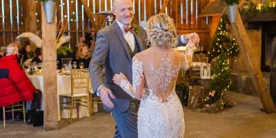 A bride and groom dancing inside a rustic wooden barn event venue with string lights and a chandelier.