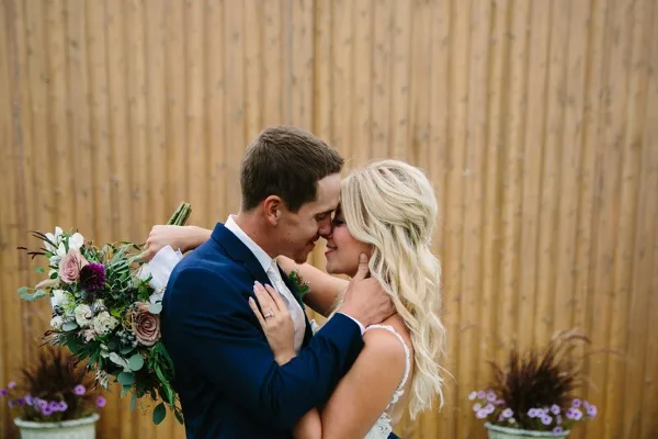 A bride and groom embracing and kissing in front of a wooden fence, with the bride holding a bouquet of flowers.