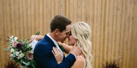 A bride and groom embracing and kissing in front of a wooden fence, with the bride holding a bouquet of flowers.