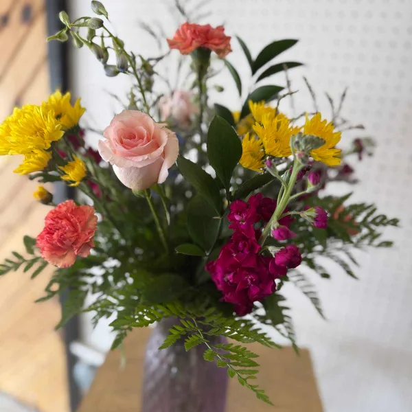A close-up of a fresh flower bouquet containing a pink rose, yellow chrysanthemums, and purple stock flowers arranged in a vase.