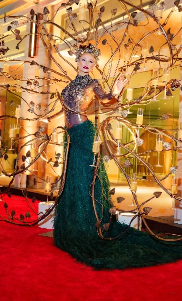 A model with dramatic makeup and an elaborate headpiece standing in front of a large, artistic metal sculpture with champagne flutes.