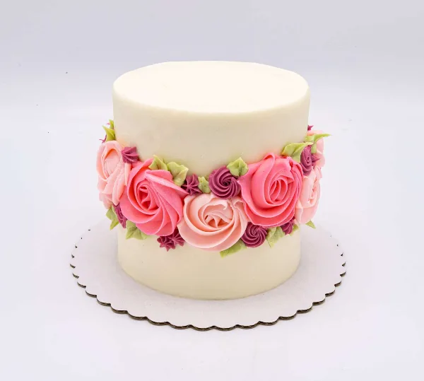 A white frosted cake decorated with a band of pink and purple buttercream roses on a scalloped cake board.