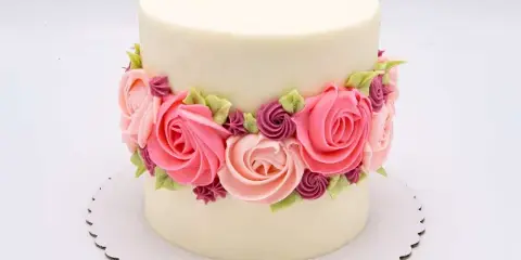 A white frosted cake decorated with a band of pink and purple buttercream roses on a scalloped cake board.