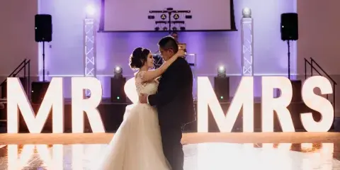 A bride and groom dancing at a wedding reception with professional stage lighting and sound equipment in the background.