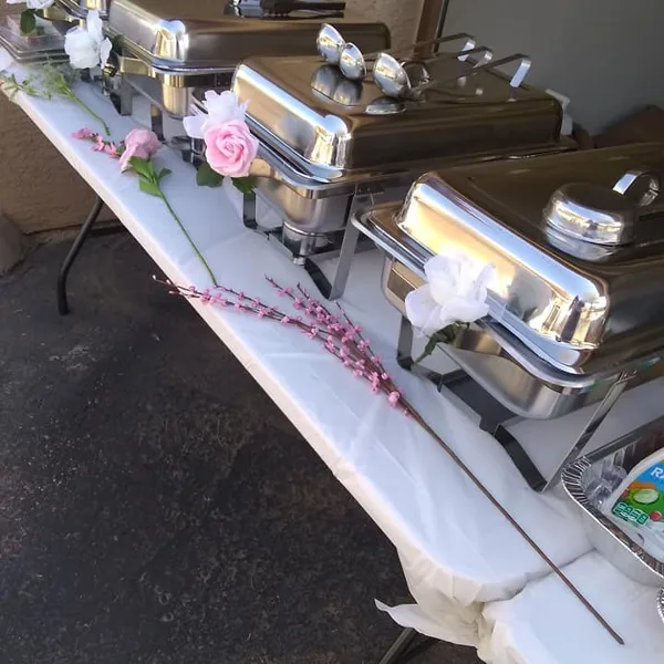 A catering buffet table with stainless steel chafing dishes, serving spoons, and floral decorations.