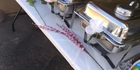 A catering buffet table with stainless steel chafing dishes, serving spoons, and floral decorations.
