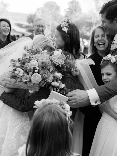 A black and white photograph of a bride and groom exchanging vows during an outdoor wedding ceremony, surrounded by smiling family members and flower girls.
