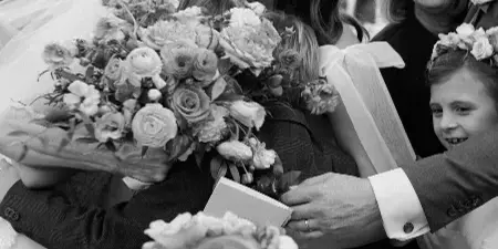 A black and white photograph of a bride and groom exchanging vows during an outdoor wedding ceremony, surrounded by smiling family members and flower girls.