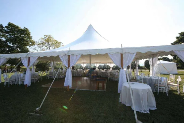 A large white event tent set up on a grassy field, furnished with round tables, white chairs, and drapery.