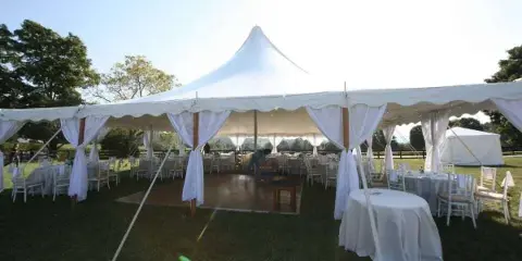 A large white event tent set up on a grassy field, furnished with round tables, white chairs, and drapery.
