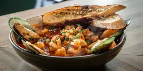 A bowl of Cioppino seafood stew with mussels, shrimp, and cod, topped with toasted baguette slices, served on a wooden table.