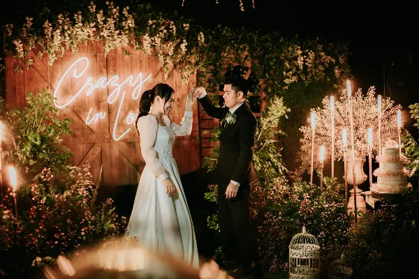 A bride and groom dancing at a wedding reception with floral decorations and a neon sign in the background.