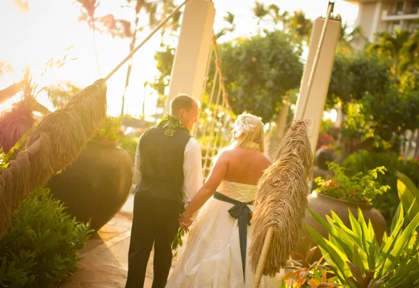 A bride and groom standing under a traditional Hawaiian wedding arch in a tropical garden setting.