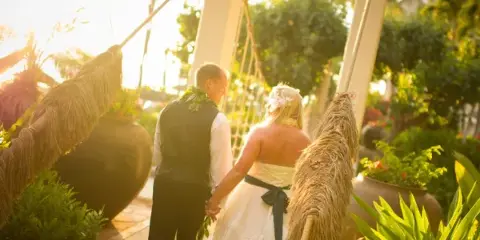 A bride and groom standing under a traditional Hawaiian wedding arch in a tropical garden setting.