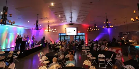 Interior view of a large event hall with a band on stage and guests seated at round tables.