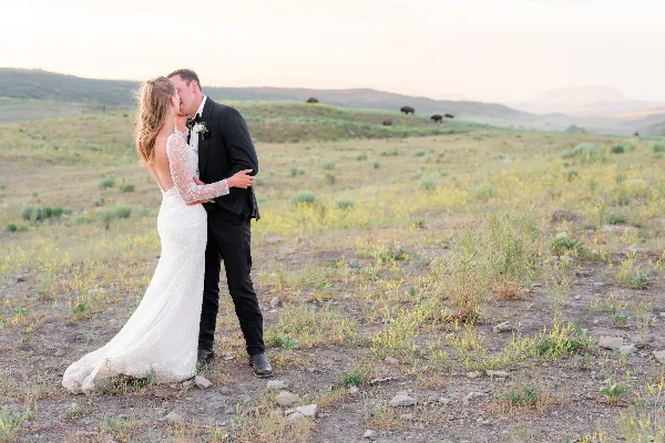 A bride and groom sharing a kiss in a grassy field with rolling hills in the background.