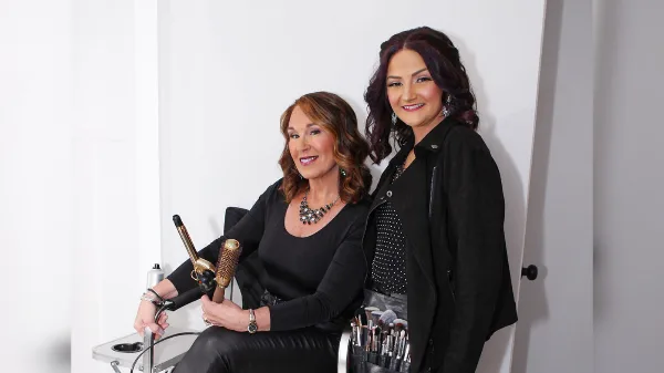 Two women posing with professional hair styling tools and makeup brushes.