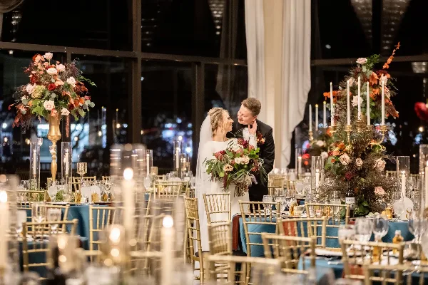 A bride and groom standing in a candlelit wedding reception hall surrounded by tall floral centerpieces and gold chiavari chairs.