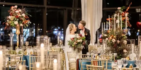 A bride and groom standing in a candlelit wedding reception hall surrounded by tall floral centerpieces and gold chiavari chairs.