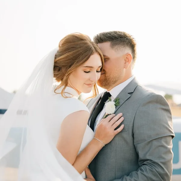 A bride in a white dress and veil embracing a groom in a grey suit with a white boutonniere.