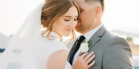 A bride in a white dress and veil embracing a groom in a grey suit with a white boutonniere.