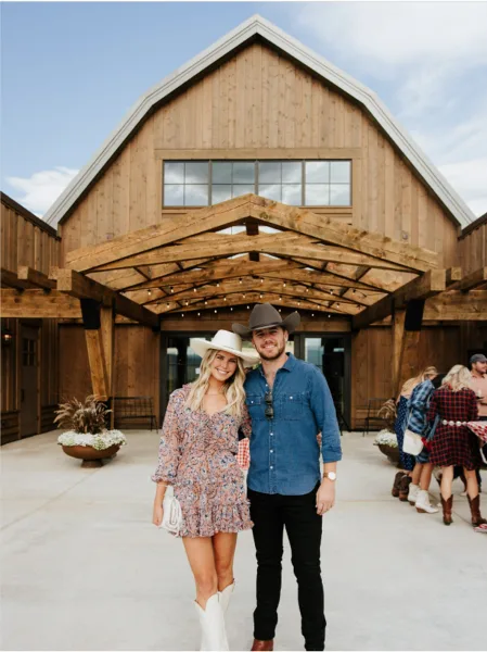 A couple posing in front of a large wooden barn venue with a covered entryway and string lights.
