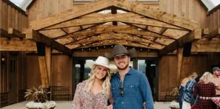 A couple posing in front of a large wooden barn venue with a covered entryway and string lights.