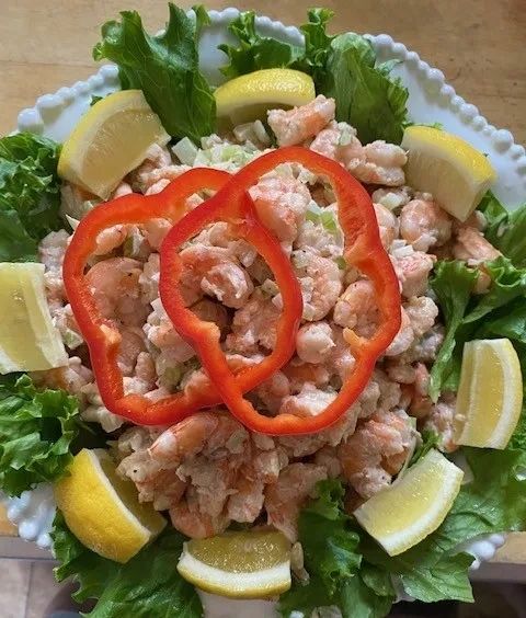A white plate filled with shrimp salad, garnished with lemon wedges, red pepper rings, and lettuce leaves.
