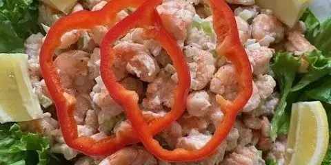 A white plate filled with shrimp salad, garnished with lemon wedges, red pepper rings, and lettuce leaves.