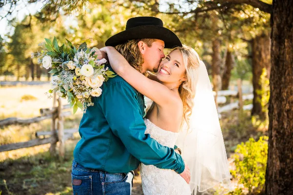 A bride and groom sharing a kiss outdoors in a wooded area, with the groom wearing a black cowboy hat and the bride holding a bouquet of white flowers.