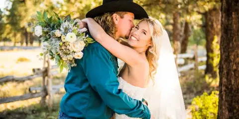 A bride and groom sharing a kiss outdoors in a wooded area, with the groom wearing a black cowboy hat and the bride holding a bouquet of white flowers.