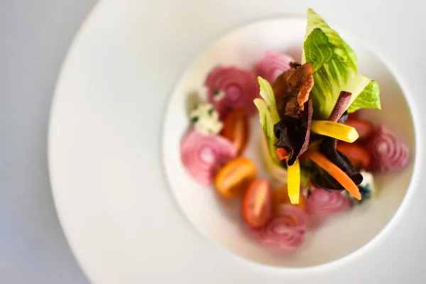 A close-up photograph of a plated salad featuring mixed greens, cherry tomatoes, and pickled red onions on a white plate.