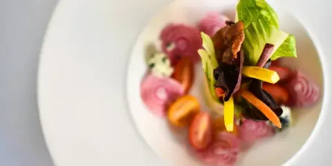 A close-up photograph of a plated salad featuring mixed greens, cherry tomatoes, and pickled red onions on a white plate.