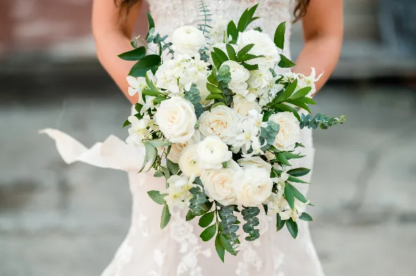 A bride in a white lace dress holding a lush bouquet of white roses, ranunculus, and greenery.