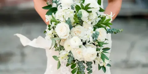 A bride in a white lace dress holding a lush bouquet of white roses, ranunculus, and greenery.