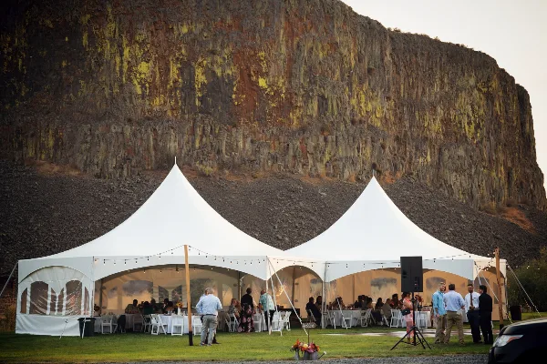 A large white event tent set up on a lawn with tables and chairs, featuring a rocky cliff face in the background.