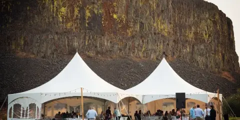 A large white event tent set up on a lawn with tables and chairs, featuring a rocky cliff face in the background.